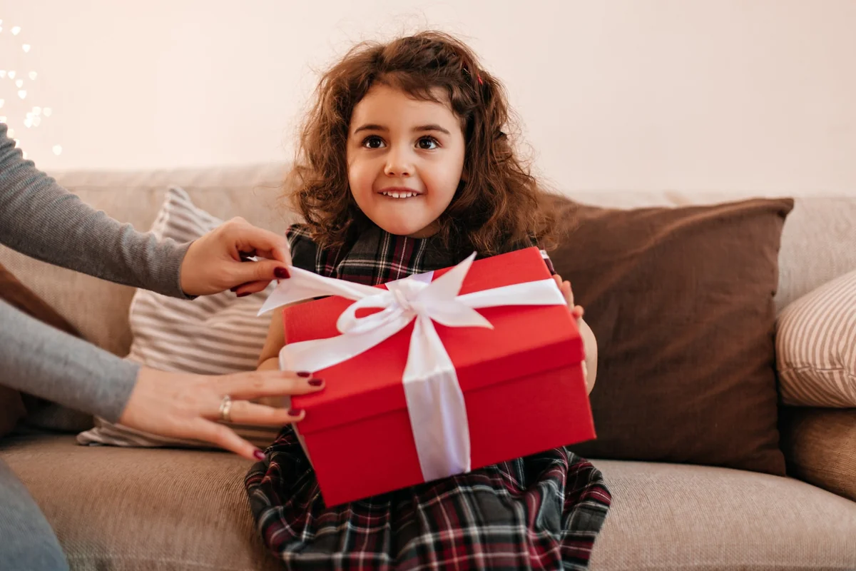 Curly-haired little girl smiling while holding a red ribbon-tied gift box, useful gifts for childrens idea