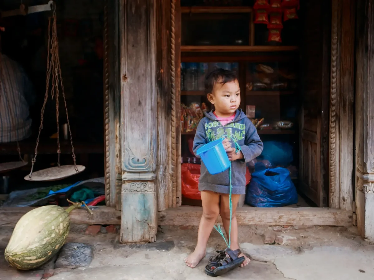 A young child stands outside a small shop holding a plastic bucket, playing with an everyday object instead of a store-bought toy