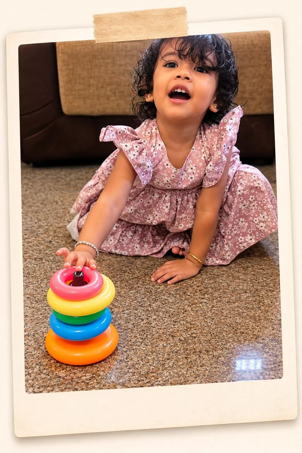 Polaroid-style photo of a toddler girl in a floral dress reaching for a rainbow ring stacker toy