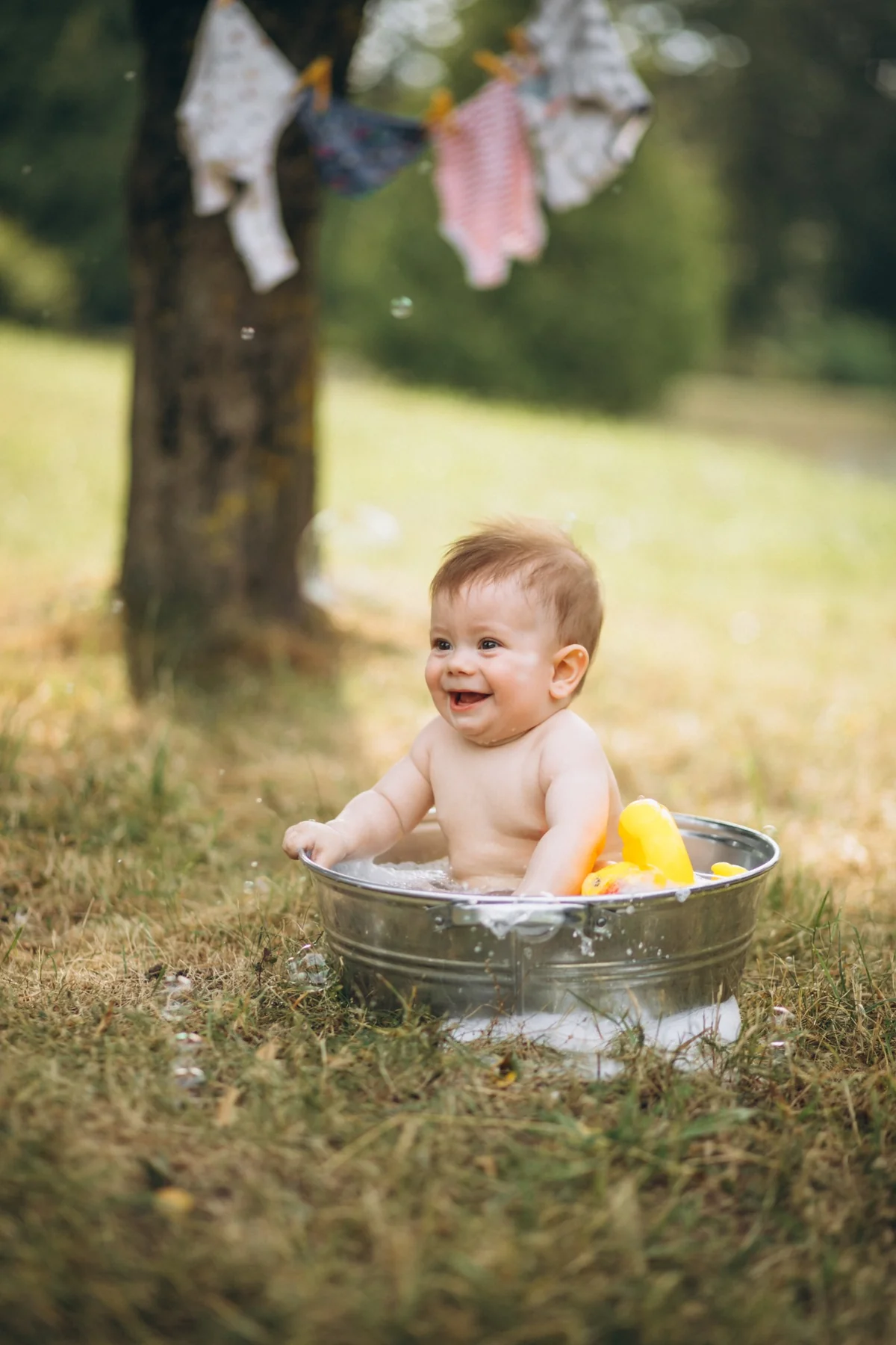 Baby laughing in an outdoor metal bathtub with a yellow duck toy floating in the water, infant bath toys outdoor bath