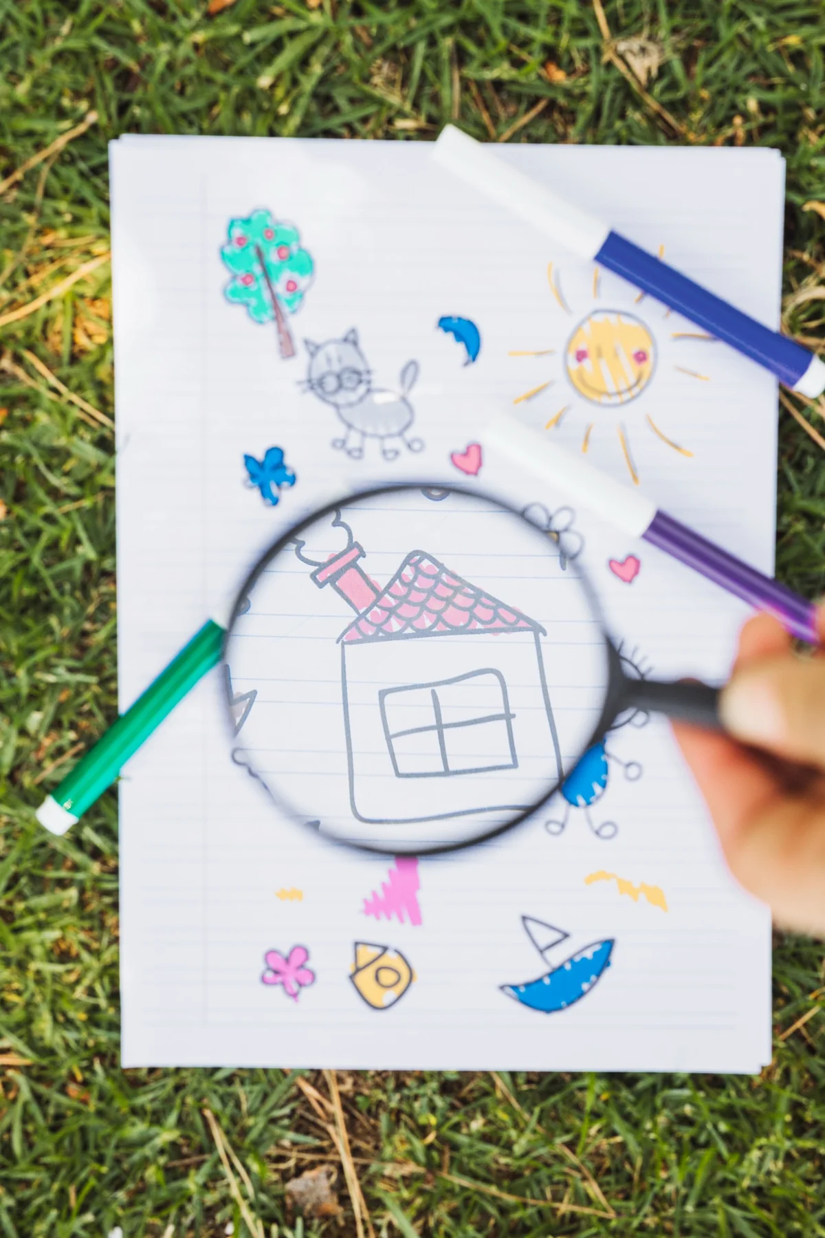 Magnifying glass over a child’s marker drawing of a house on lined paper, with colored markers lying on the grass