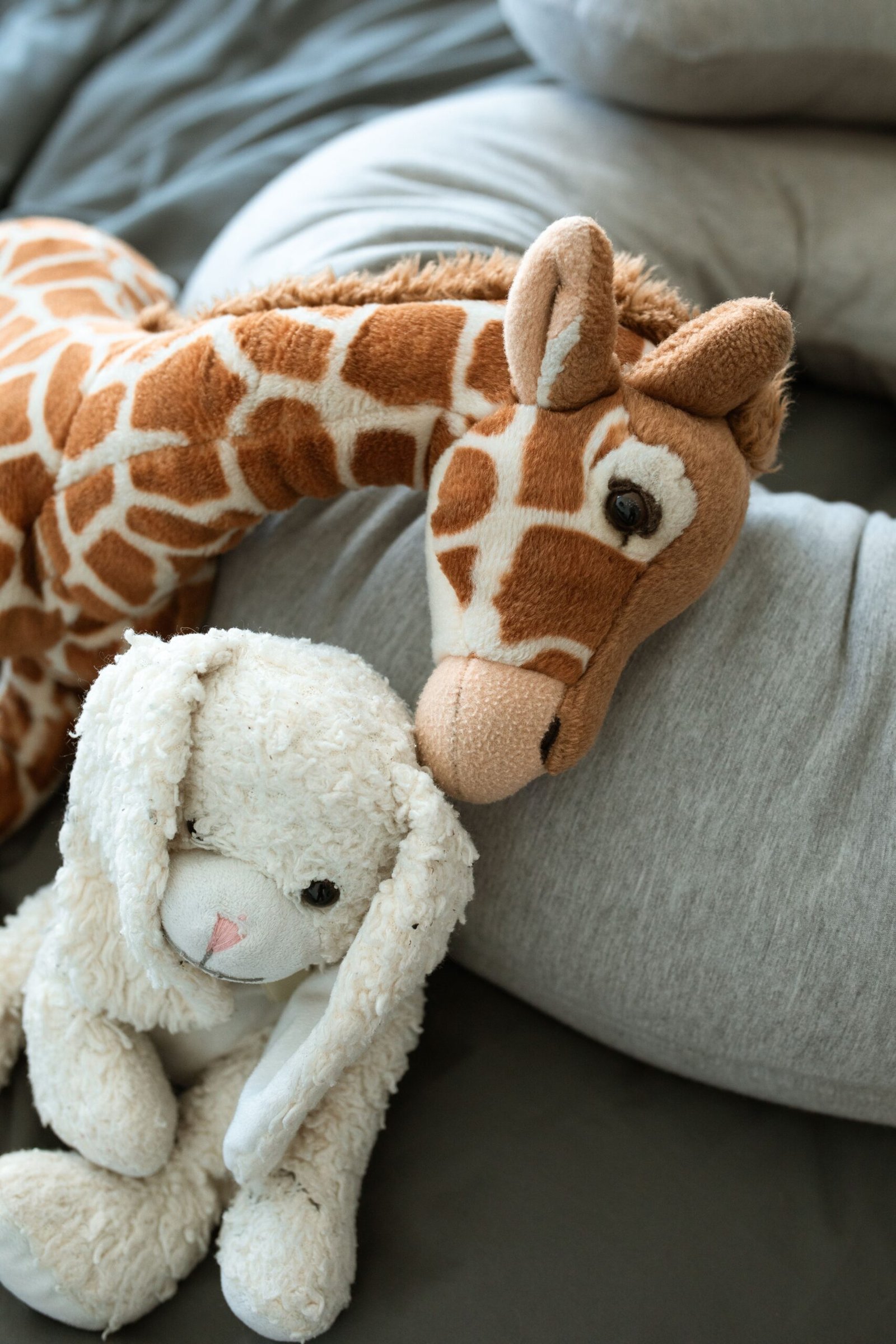 Plush giraffe resting its head against a white fluffy bunny on grey bedding, soft toys for newborn