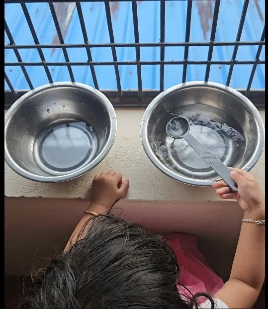 Top view of a child at a window ledge with two steel bowls of water and a small plastic scoop, fine motor activities